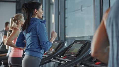 Fit Athletic Woman Running on the Treadmill, Doing Her Fitness Exercise. Muscular Women and Men Actively Training in the Modern Gym. Sports People Workout in Fitness Center. Side View Slow Motion