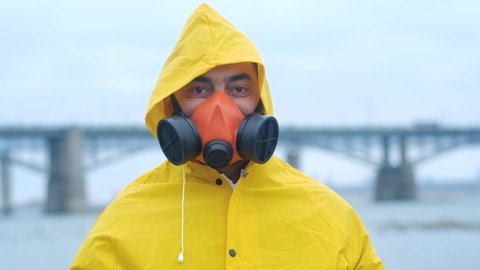 chief ecologist expert in yellow raincoat removes red mask and inhales fresh air. Specialist stands on city river on background of bridge. Care and preservation of environment. Outside water analysis