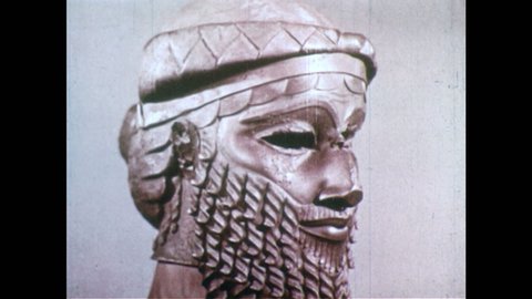 1950s: Sumerian statue, man with beard. Statue of bald person wearing skirt with writing on back. Person uses pointed wooden stick to write on clay tablet.