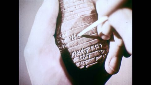 1950s: Hands hold clay tablet with Sumerian cuneiform writing. Round barrel shaped clay object. Hand holds clay tablet, uses pointed stick to count lines in symbols.