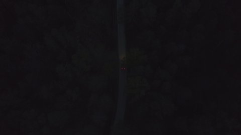 Aerial front view black business sedan car driving on dark countryside asphalt road among summer forest at night. View from drone car with headlights riding on night road through dark trees