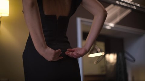 A girl in a black dress elegantly fastens a zipper on her back before a meeting in a dark room in the hotel