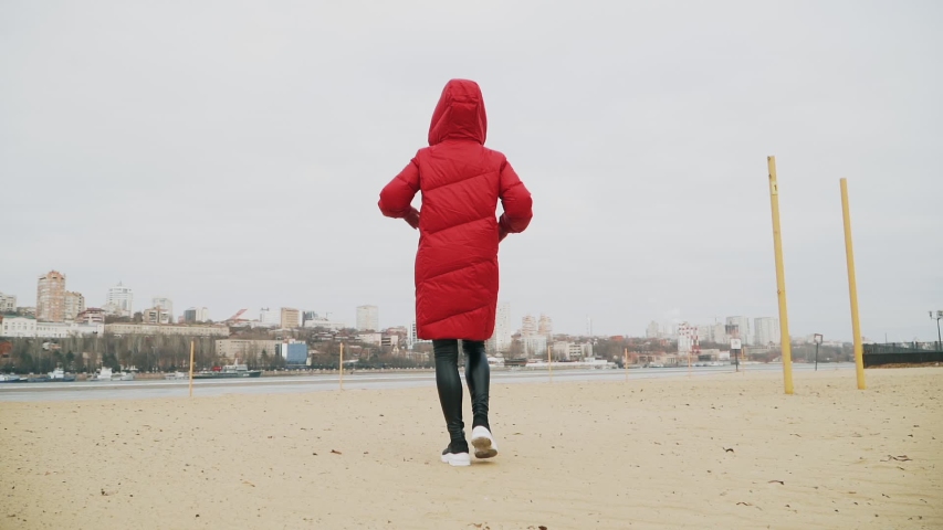 A young girl warmly dressed in a red down jacket and gray hat is walking along the river. Cold. Strong wind. Against the background of the city. Slow motion.