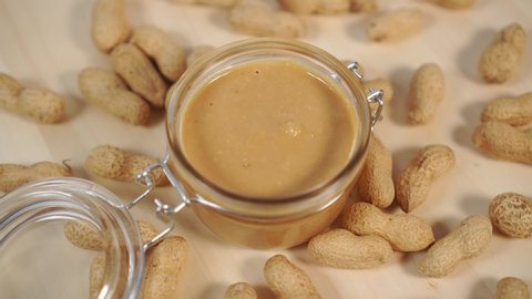 Peanut butter in a glass jar with a metal fastener on a wooden table close-up with a bunch of nuts. Rotation. Healthy Organic Breakfast