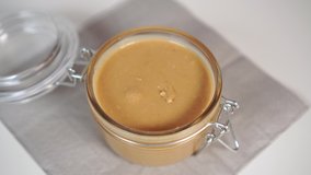 A girl takes a knife with juicy homemade peanut butter in a glass jar with a metal clip on a gray napkin. Making Healthy Organic Breakfast