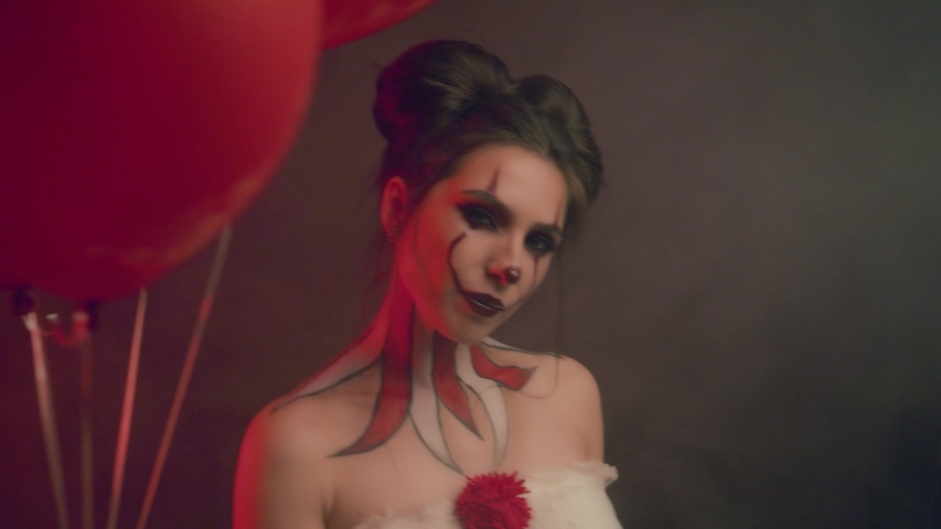 happy young woman in costume makeup crazy clown smiling laughing dancing look. Holds in hand red balloons. positive emotions. Backdrop dark gothic studio smoke. animator children