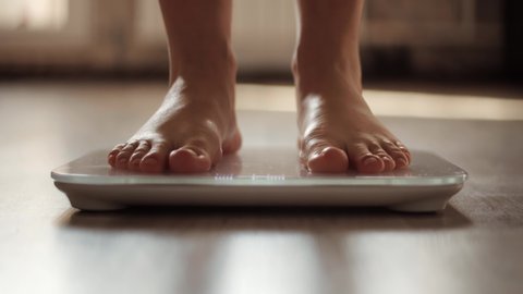 Woman On Scales Measure Weight. Girl Legs Step On Bathroom Scale. Diet Female Feet Standing Weighing Scales On Room. Human Barefoot Measuring Body Fat Overweight. Slim Woman Checking BMI Weight Loss
