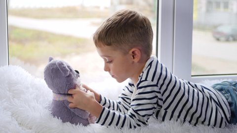 A cute little boy shares secrets with a Teddy bear toy while lying on the sill on a cloudy day. Children's dreams and emotions.