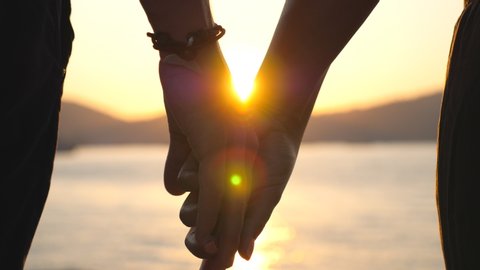 Male and female hands holding each other at sunset. Young couple standing on seashore and enjoying spending time together. Romantic moment between two lovers. Concept of loving or happiness. Slow mo