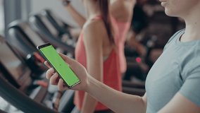 Athletic Woman Uses Green Mock-up Screen Smartphone while Running on the Treadmill in a Gym. Sports Female Posts on Social Media, Takes Pictures, Watches Videos while Exercising in the Fitness Club