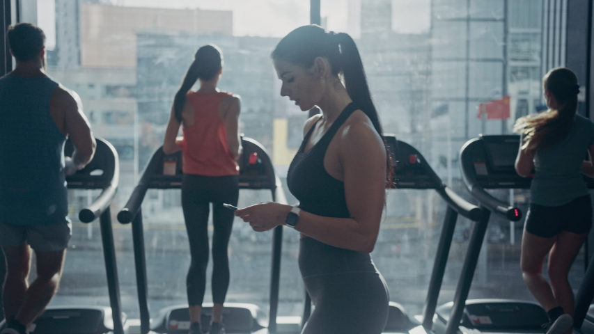 Beautiful Athletic Woman Uses Smartphone Walks Through Fitness Club Gym. Using Social Media, Posting Pictures, Communicating, Checking Email. In the Background Sports People Running on Treadmills Royalty-Free Stock Footage #1041686002
