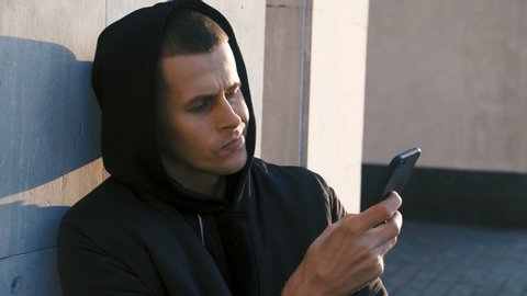 Portrait of young man gangster drug dealer in the hood uses a smartphone outdoor urban background