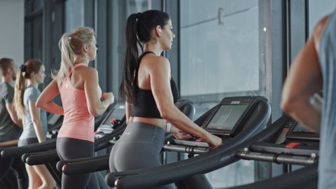 Beautiful Athletic Sports Woman Puts on Wireless Headphones, Turns on Podcast / Music Playlist with Smartphone and Starts Running on a Treadmill in the Gym. Slow Motion Side View