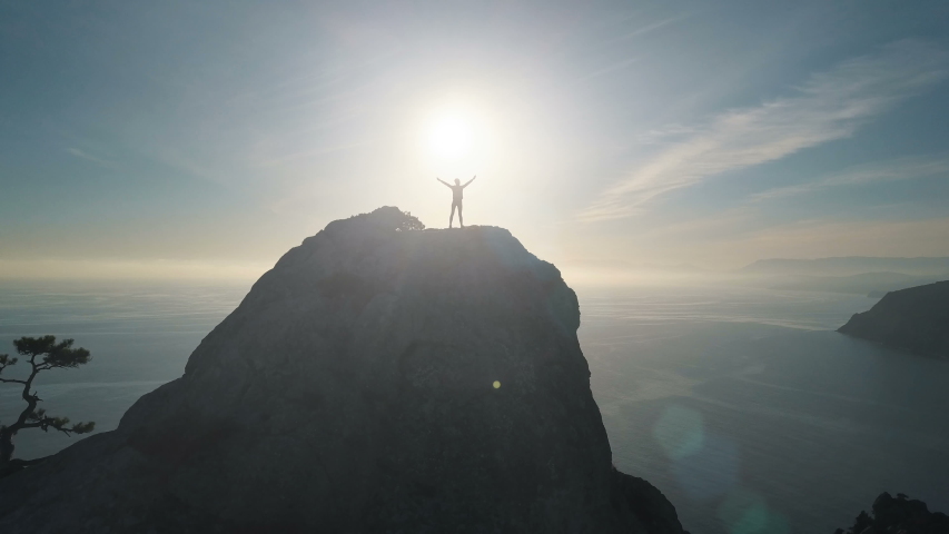 Aerial silhouette of a young woman climbing to the top of a mountain against the sea at sunset. Lady on the summit in beautiful scenery raises her hands up. Royalty-Free Stock Footage #1041688321