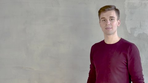 Portrait of young man short cut burgundy cardigan standing against gray wall looking to the left demonstrates presentation points with his hand at 4 positions turns his head to camera smiling close up