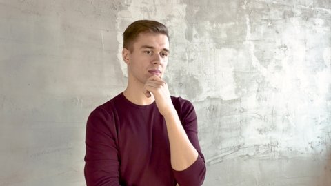 Young man in burgundy jumper stands against gray wall thinks about solution to problem rubs his chin raises his finger up smiling Free space for inscriptions on the right Middle shot