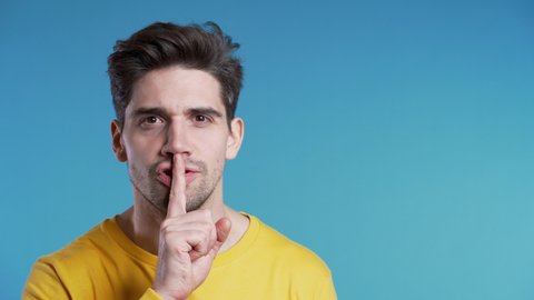 European handsome man holding finger on his lips over blue background. Gesture of shhh, secret, silence. Copy space