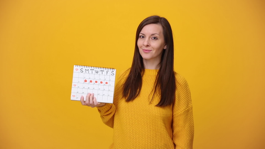 Young woman in sweater hold in hand female periods calendar for checking menstruation days show thumb up isolated over yellow background in studio. Medical healthcare gynecological lifestyle concept.