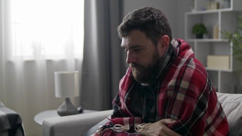 A man coughs heavily while sitting in a plaid on a couch in the living room