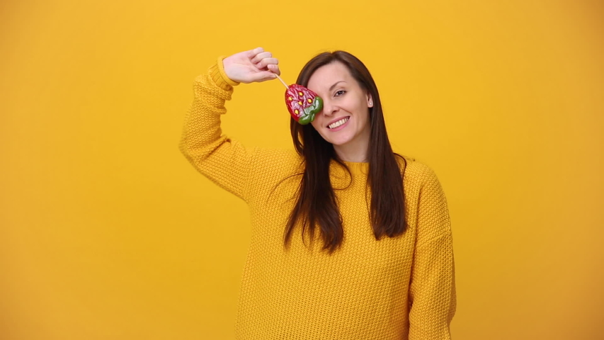 Smiling beautiful brunette young woman in bright yellow sweater shirt posing isolated over yellow orange background in studio. People sincere emotions lifestyle concept. Hold lollipop candy strawberry