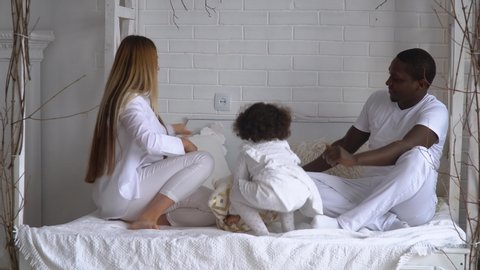 Happy mixed ethnicity family mom, african dad and little kid daughter having fun pillow fight on bed