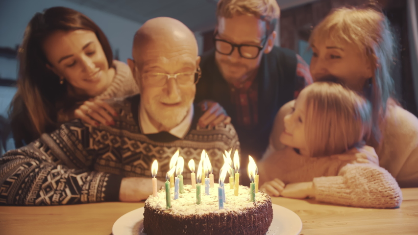 Celebration and family concept - happy grandfather blowing candles on birthday cake at dinner party surrounded by his large family. Royalty-Free Stock Footage #1041705808