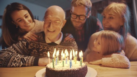 Celebration and family concept - happy grandfather blowing candles on birthday cake at dinner party surrounded by his large family.