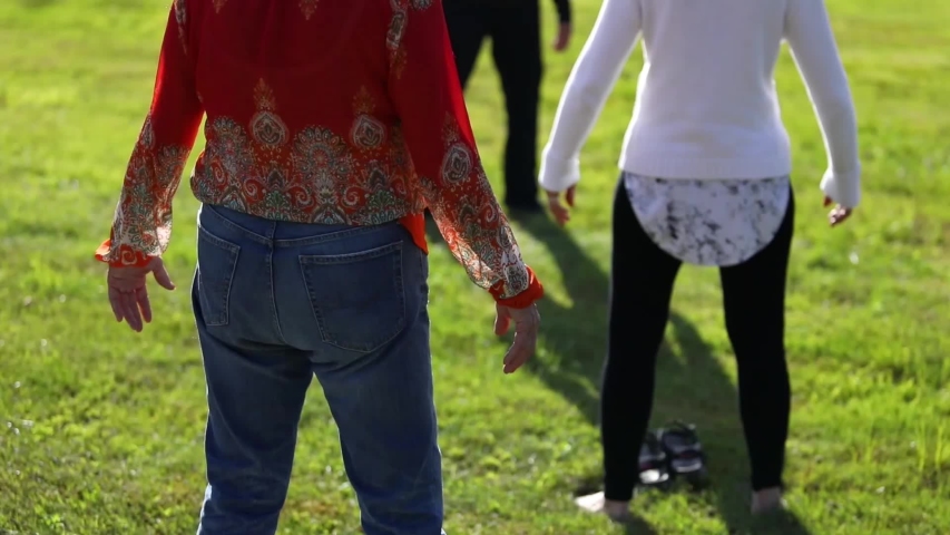 Morning exercise class in the park. Students seen from the back doing deep breathing with the instructor in slow motion and selective focus.