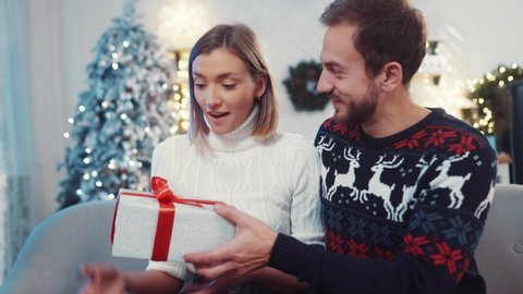 Young happy man closing eyes of his wife and giving a lovely christmas present. Euphoric young woman receiving pleasant gift hugging her lover celebrating winter holidays.