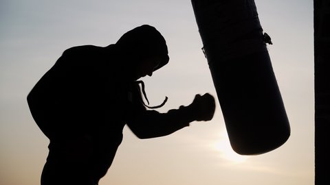 Boxer beats punching bag on sunset background. Silhouette motivation sport boxing. Epic video. Martial arts. Training in the fresh air.