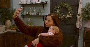 Caucasian mother holding her child on hands and taking selfie in kitchen with Christmas decorations. RAW Graded footage 4K slow motion 50fps