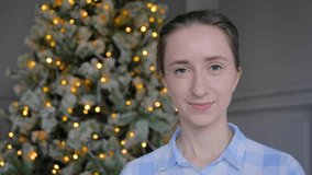 Slow motion: video portrait of positive hipster, student or entrepreneur woman in plaid shirt smiling and looking at camera in room with Christmas tree. Garland light illumination. Holiday concept