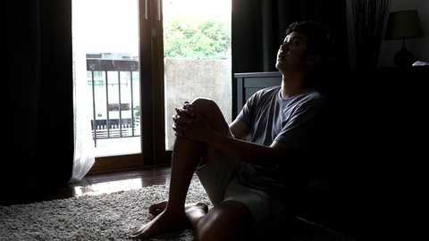 HD Slow motion young asian depressed sad man sitting on bedroom floor near balcony at sunset. Distraught upset man sitting alone with hugging the knees and looking away. Mental health care concept.