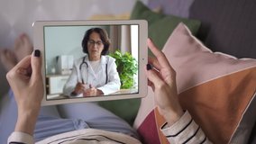 Online video call via tablet of female doctor in white lab coat explaining to her patient behind the screen. sick woman lying on sofa listening. concept medicine, healthcare, internet