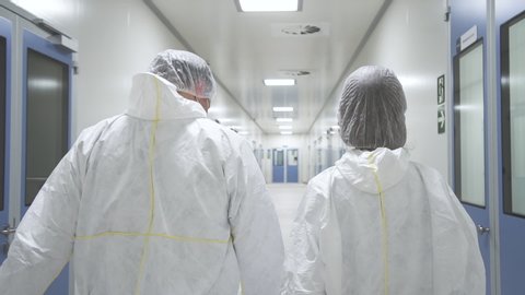 American man and woman worker walk and having talk in pharmaceutical plant. Two people employee discussing work ideas in interior of medical company. Back view of young staff wearing white protective