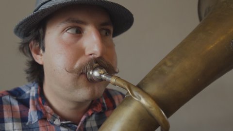 Cheerful young man with a funny mustache and a hat playing on the big tuba. Close up
