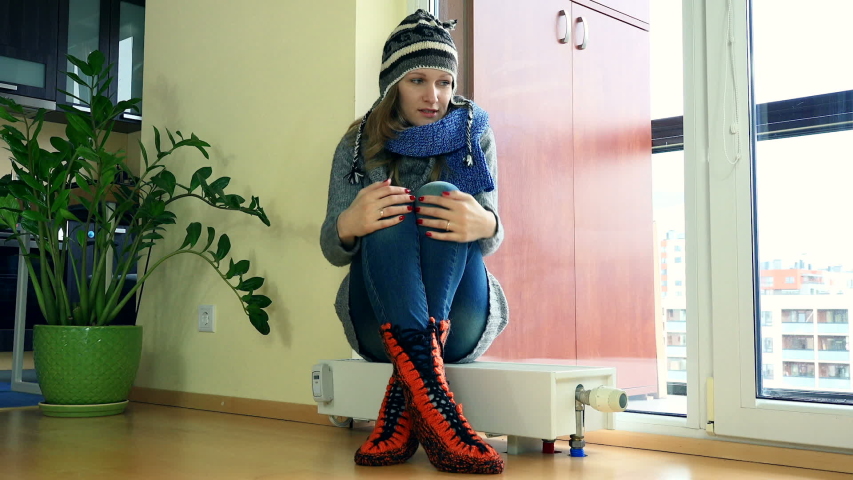 Cheerful girl strip warm clothes sitting on heater near balcony window. Beginning of central heating at flat house.
