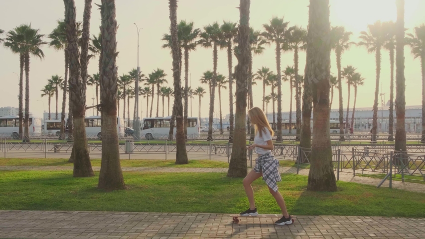 Riding a Longboard on a Sunny Day. A beautiful Blonde Woman on a skateboard in modern, Trendy Clothes rides through the Park past the palm trees. The tracking shot. 4K. Slow motion, side view.