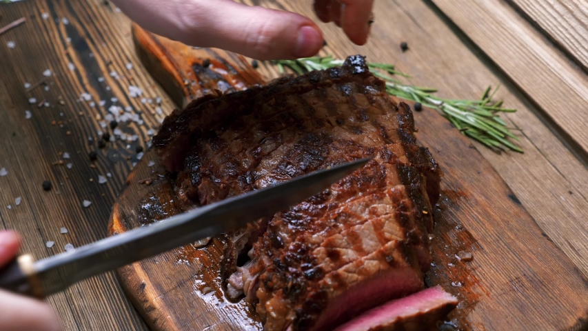 Man Hand with Vintage Knife Checking of Chopped Grilled Beef Steak on Rustic Cutting Board on Wooden Background. Juicy Sliced Rib Eye with Salt And Rosemary on Table. Concept of Delicious Meat Food | Shutterstock HD Video #1041786181