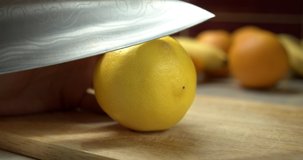 
Damascus knife cuts juicy lemon. Juice flows onto the cutting board. Bananas, oranges and lemons on the background. Yellow color. Slow zoom in.