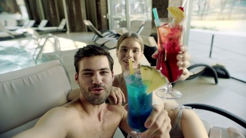 Happy couple making selfie photo on mobile phone at poolside. Pov of smiling guy and girl drinking cocktails at loungers. Portrait of sexy couple clinking glasses for self portrait at poolside.