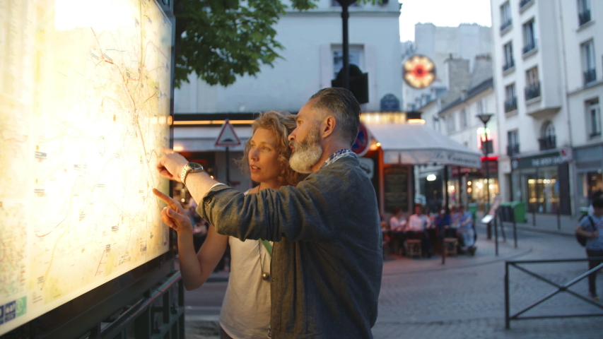 A middle aged couple look over a public transportation map in downtown Paris, France. Two tourists enjoy a romantic evening out together on a date in Europe. Travel, tourists, commuting, hand-held.