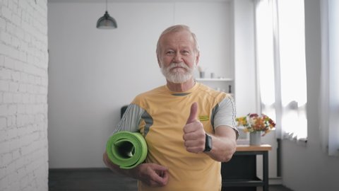 Portrait of a handsome satisfied old man in great shape with a yoga mat in his hands and a smart watch, after playing sports to maintain his health and showing a thumbs up gesture while standing