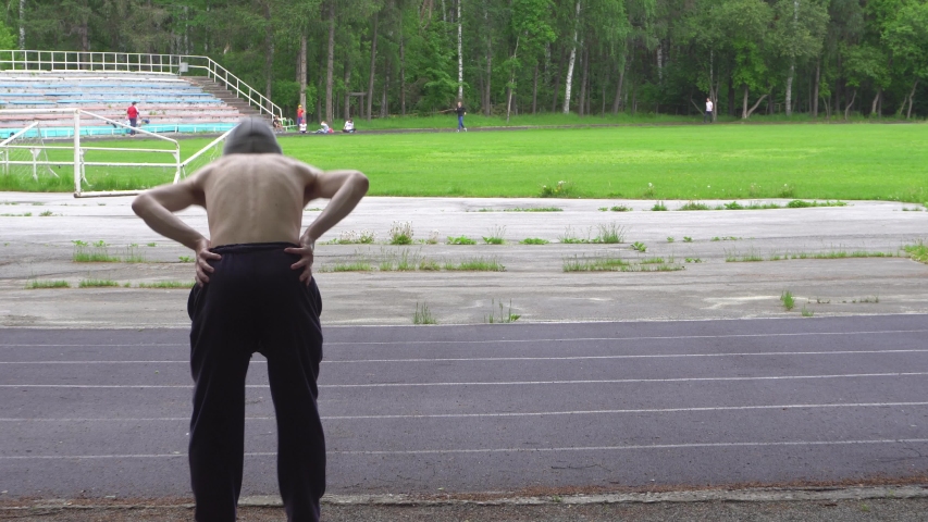 An elderly sporty man warming up before running in the park at the stadium. A view from the back. 4k footage.