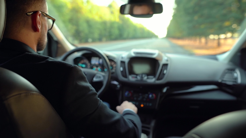 Formally dressed bearded man in glasses driving a car in sunny weather. View from the back seat of a car