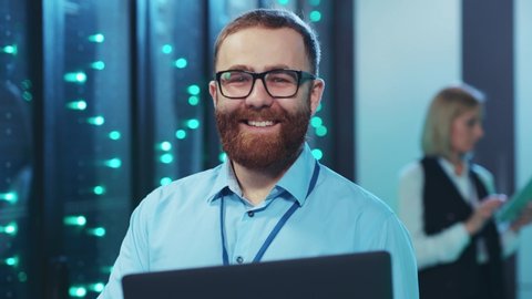 Handsome bearded IT administrator with laptop smiling cheerfully on camera coworking in cyber security cloud computing webhosting at data center.
