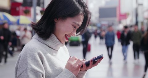 A Chinese young woman is looking down at her mobile phone happily.A woman is scannning her cellphone cheerfully in a busy road.An Asian female is browsing her smart phone with her head down outdoors.