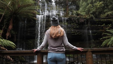 Young Blonde Traveller Looks up at Russel Falls in Tasmania Tiered Cascading Waterfalls in a Fern Jungle Forest, Amazing Fern Jungle View