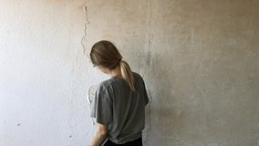 Video 4K. Young hipster girl makes repairs in the flat. Clean old plaster on the walls before paint into new white color. Works with putty knife . 