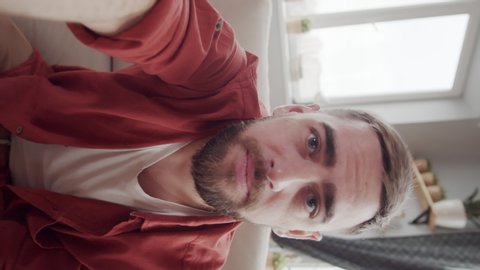 Waist-up handheld shot with vertical screen orientation of casually dressed bearded young Caucasian man with outstretched hand sitting at home and having lively conversation on video call